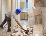 Obrero con casco azul trabajando en la demolición de un tabique, rodeado de escombros.