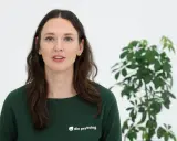 En kvinna med mörkt hår och blå ögon, klädd i en grön tröja med texten "din psykolog", talar. Bredvid henne står en grön växt. Hon ger dig verktyg att hantera sorg.