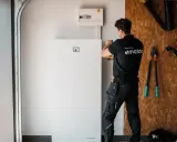 Tekniker installerar ett stort, vitt solpaneler batteri i ett garage. Verktyg hänger på väggen bakom.