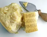 Ein Stück Parmesan mit eingeprägtem Schriftzug und ein Käsemesser. Aus was besteht Parmesan? Aus Milch, Lab und Salz.
