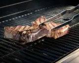 Zwei saftige Cowboy-Steaks werden mit einer Zange auf einem heißen Grill gewendet. Rauch steigt auf, während die Flammen darunter lodern.