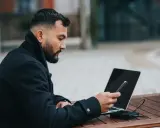 Mężczyzna z brodą i laptopem na stoliku, trzymający telefon. Zastanawia się, co zrobić, żeby ktoś odpisał.