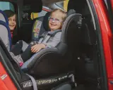 Dwie uśmiechnięte dziewczynki w fotelikach Britax Romer 0-18 kg w czerwonym samochodzie.