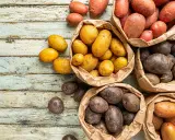 Une variété de pommes de terre, allant du jaune doré au rouge et au violet foncé, est présentée dans des sacs en papier sur une table en bois vieilli.