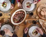 Un repas du terroir avec viande, saucisses, fromages et pain sur une table en bois. Des mains servent et dégustent.
