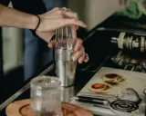 Ręce mieszają składniki w shakerze, przygotowując drinki na ciepło. Na blacie leży butelka whiskey i przypalone plasterki jabłka.