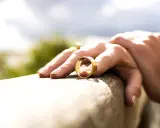 Dłoń z obrączką na drewnianej powierzchni, na tle nieba i zieleni.