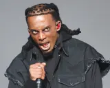 Playboi Carti z mikrofonem, w czarnej kurtce i z charakterystycznymi warkoczami. Jego ekspresja sugeruje energię znaną z jego playboi carti utworów.
