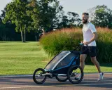 Ein Mann joggt mit Kinderwagen durch eine grüne Parklandschaft.