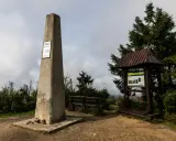 Kamienny obelisk na szczycie Turbacz w Gorcach, obok drewniana wiata z tablicą informacyjną i ławką.