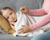 Dziecko leży pod kocem, mama mierzy mu temperaturę. Wysoka gorączka u dziecka bez innych objawów martwi.