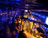 Lebhafte Nacht in einem der Prager Clubs, beleuchtet von blauem und gelbem Licht. Menschen stehen an der Bar und unterhalten sich.