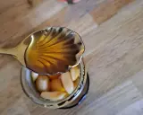 Une cuillère dorée verse du miel ambré sur des gousses d'ail confites dans un bocal. Un délice d'ail au miel.
