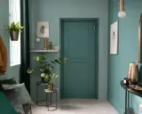 Entrada decorada con pared verde azulado, puerta verde, perchero con abrigo y bolso, plantas y espejo.