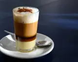 Ein geschichteter Kaffee con leche in einem Glas, mit einer Löffel auf einer Untertasse.