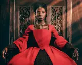 Jodie Turner-Smith, con su mirada regia, se sienta en un trono de madera tallada, vestida con un opulento traje rojo.