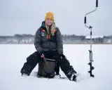 Kvinna ler och njuter av att fiska på isen. Bredvid henne står en isborr redo för användning.