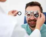 Mężczyzna uśmiecha się podczas badania wzroku w domu. Optometrysta wymienia soczewki w oprawce próbnej.