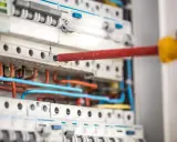 Ręka z czerwonym śrubokrętem pracuje przy instalacji niskoprądowej. Widać bezpieczniki i kolorowe przewody.