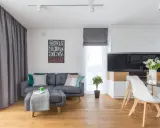 Mały salon inspiracje: szara kanapa, stolik z pufą, nowoczesna kuchnia i jadalnia. Drewniana podłoga dodaje ciepła.