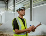 Un ingeniero de estructuras con casco y chaleco reflectante anota datos en una libreta, supervisando la construcción.