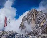 Austriacka flaga na szczycie góry, symbolizująca pracę w Austrii. W tle skaliste zbocza i chmury.