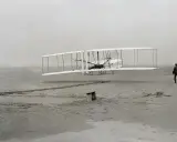 Der Wright Flyer hebt vom sandigen Boden ab, ein Mann beobachtet den historischen Flug.