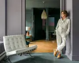 A woman in a cream jumpsuit leans against a wall in the robin rice gallery, with a Barcelona chair and abstract art visible.