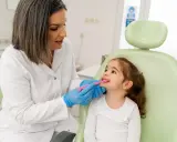 Dentystka w białym fartuchu i niebieskich rękawiczkach pokazuje małej dziewczynce, jak prawidłowo szczotkować zęby. To przykład troski o zdrowie jamy ustnej w ramach pedodoncji.