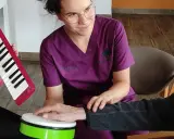 Terapia dźwiękiem i rytmem: muzykoterapia dla seniorów. Terapeuta wspiera dłoń pacjenta na bębenku, podczas gdy inny uczestnik gra na keyboardzie.