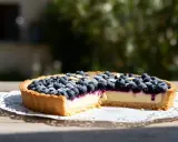 Une délicieuse tarte myrtille amande, garnie de myrtilles fraîches et d'amandes effilées, sur une table en bois ensoleillée.