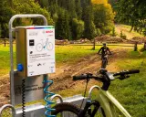 Stacje ładowania rowerów elektrycznych w Piwnicznej-Zdroju. W tle rowerzystka na szlaku.