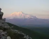 Elbrus, najwyższy szczyt Rosji, lśni w blasku zachodzącego słońca. W oddali widać zielone wzgórza i skaliste zbocze.