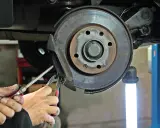 Mechanik wymienia linkę hamulca ręcznego, używając narzędzi do demontażu zacisku hamulcowego przy tarczy.