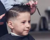 Chłopiec z zaciekawieniem obserwuje, jak powstają modne fryzury dla chłopców. Fryzjerka precyzyjnie modeluje jego włosy.