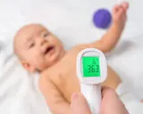 Jak mierzyć temperaturę termometrem elektronicznym? Dziecko leży na białym tle, a termometr pokazuje 36.3°C.