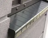 Szary, metalowy parapet zewnętrzny z widocznym uszczelnieniem. Detale mocowania okna i faktura muru.