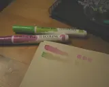 Dwa pisaki Ecoline (zielony i fioletowy) na drewnianym stole, obok próbki kolorów na papierze.