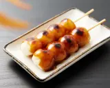 Brochettes de dango grillés, nappées de sauce sucrée, sur une assiette blanche.