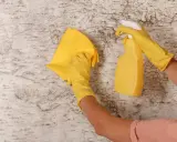 Manos con guantes amarillos limpiando paredes con un paño y spray.