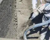 Un trabajador aplica hormigón proyectado a una pared con una manguera. El material salpica al ser aplicado.