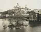 Widok na miasto z rzeką i kościołem. W oddali widać potężną budowlę sakralną z kopułami. Na pierwszym planie rzeka z wysepką i drzewami. Po prawej stronie drewniane budynki nad wodą. Ktoś pisze na Berdyczów.