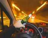 Ręka trzyma butelkę piwa za kierownicą. Po ilu można jechać autem po alkoholu? Nigdy!