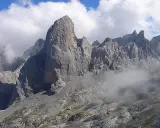 Majestatyczny **Naranjo de Bulnes** w Picos de Europa, otoczony mgłą i chmurami, z małym schroniskiem u podnóża.