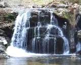 Cascada en el **valle de Cabuérniga**, con agua cayendo en varios niveles sobre rocas oscuras y musgosas, formando un remanso tranquilo.