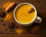 Une tasse de lait au curcuma, avec une bâton de cannelle, sur fond de bois. Autour, des épices : curcuma, anis étoilé, poivre noir.