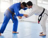 Dwóch mężczyzn w kimonach do BJJ, jeden w niebieskim z brązowym pasem, drugi w białym z czarnym pasem, przygotowuje się do walki.