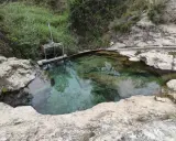 Aguas cristalinas en los baños de Somogil, rodeadas de rocas y vegetación. Una compuerta metálica controla el flujo del agua.
