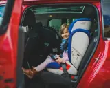 Uśmiechnięta dziewczynka w foteliku samochodowym Britax Römer w czerwonym aucie.