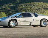 Silberner Isdera Imperator 108i mit markanten Lufteinlässen und futuristischem Design, geparkt vor einer grünen Hügellandschaft.